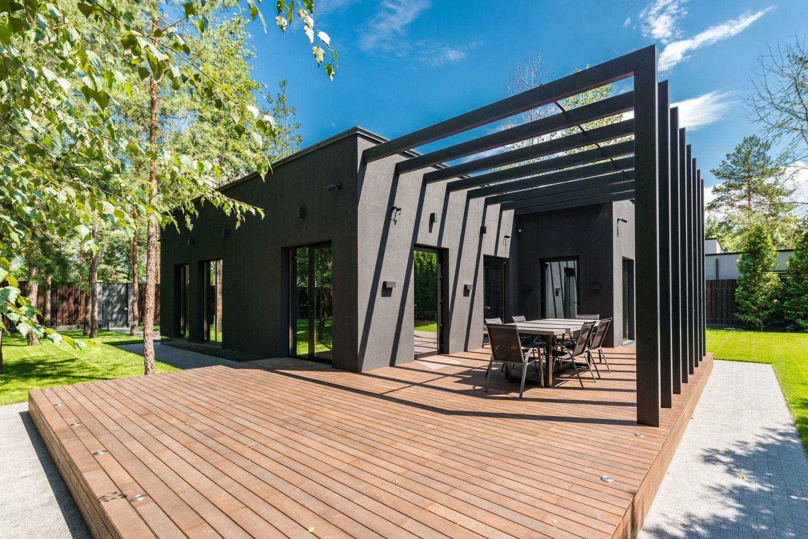 Bred terrasse af trælister foran et mørkt hus med sort pergola